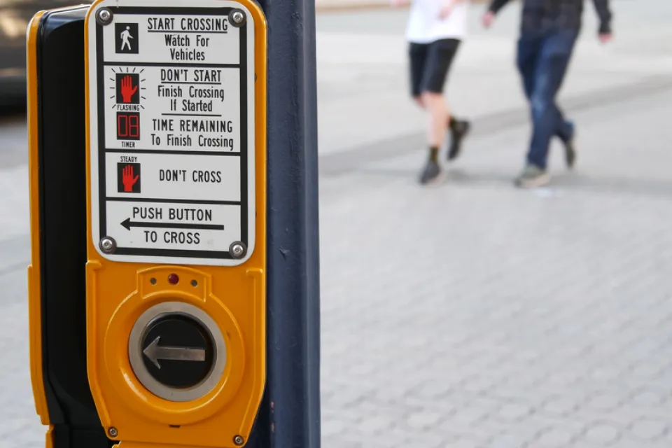 A crosswalk activation button is shown in the foreground with two pedestrians show blurred while crossing the street in the background.